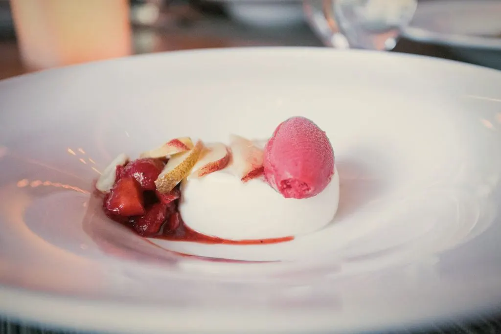 Panna cotta na głębokim białym talerzu z truskawkami, plasterkami brzoskwini i kulką różowego sorbetu
