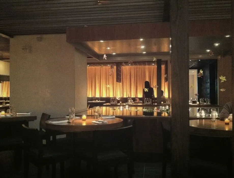 Ciemna sala restauracji z drewnianymi stołami, w głębi bar podświetlony ciepłym światłem zza zasłon